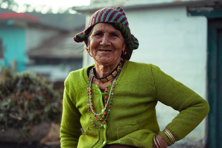 A joyful elderly woman wearing a green knitted sweater and colorful beanie smiles warmly outdoors.
