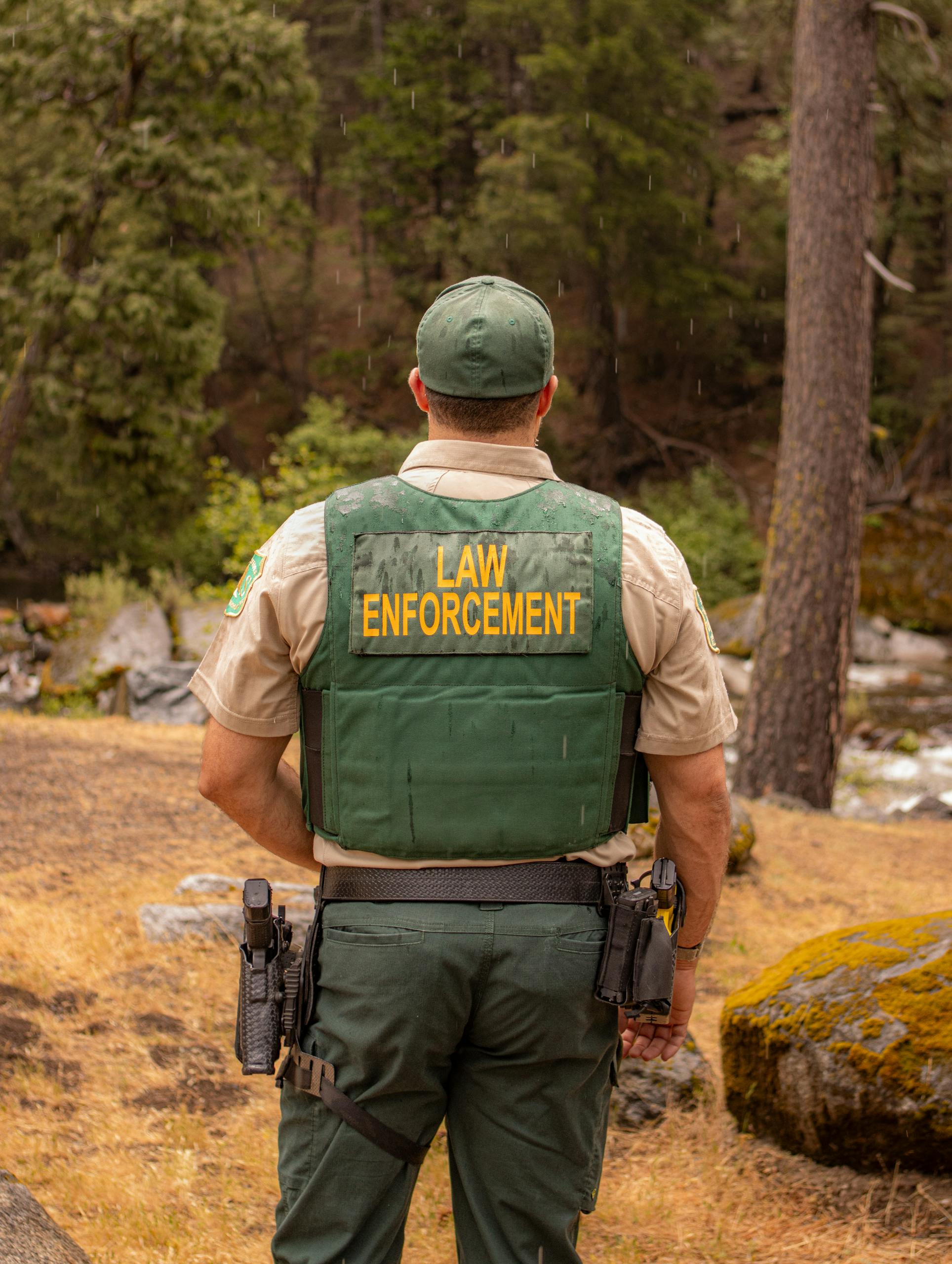 A law enforcement officer standing in a forest, viewed from behind.