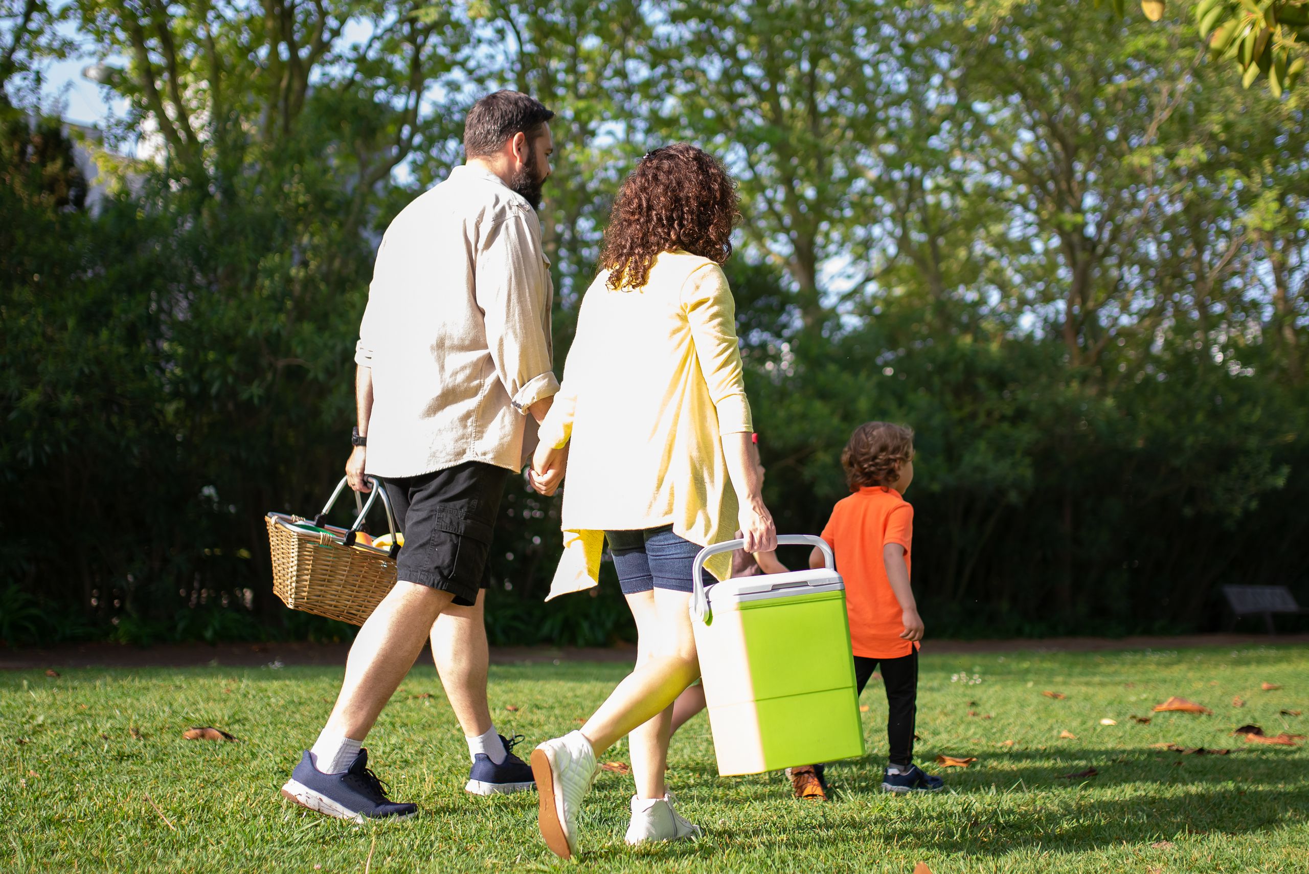 A family with a child carrying picnic essentials walking in a sunny park.