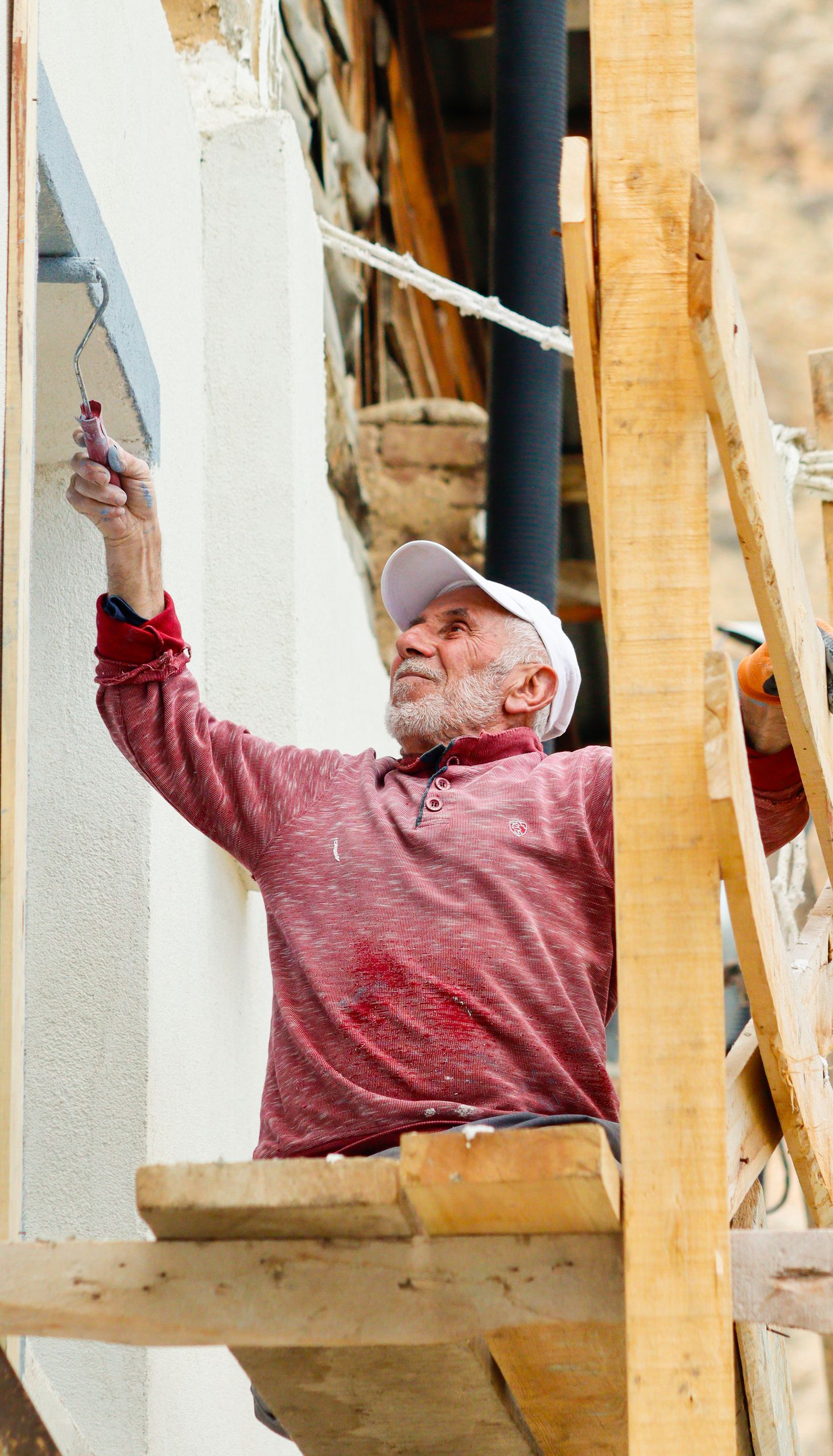 Elderly man wearing a cap paints an exterior wall while seated on scaffolding.